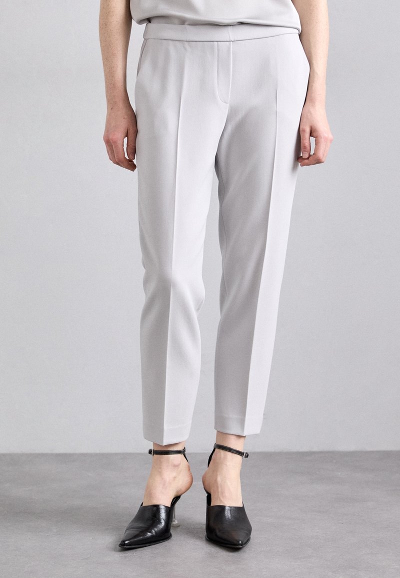 Personne portant un pantalon gris clair sur mesure longueur cheville avec des talons noirs à bout pointu et brides à la cheville, debout sur un sol gris.