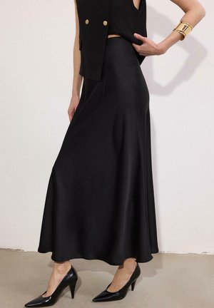 Kobieta ubrana w czarną bezrękawnikową kamizelkę z złotymi guzikami, czarną satynową długą spódnicę, czarne szpilki oraz szeroką złotą bransoletkę na lewym nadgarstku.