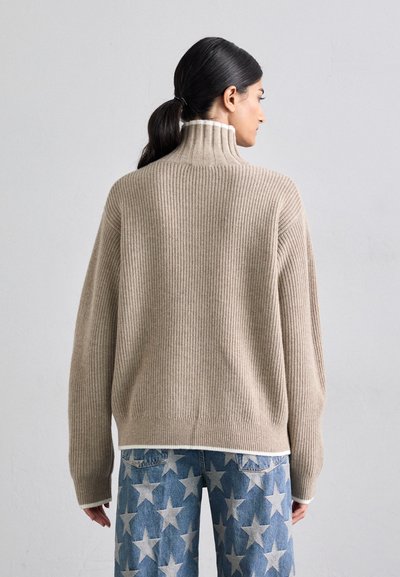 Beige Rippstrickpullover mit hohem Stehkragen und lockerer Passform, kombiniert mit blauen Sternenmuster-Hosen mit einem verwaschenen Look.