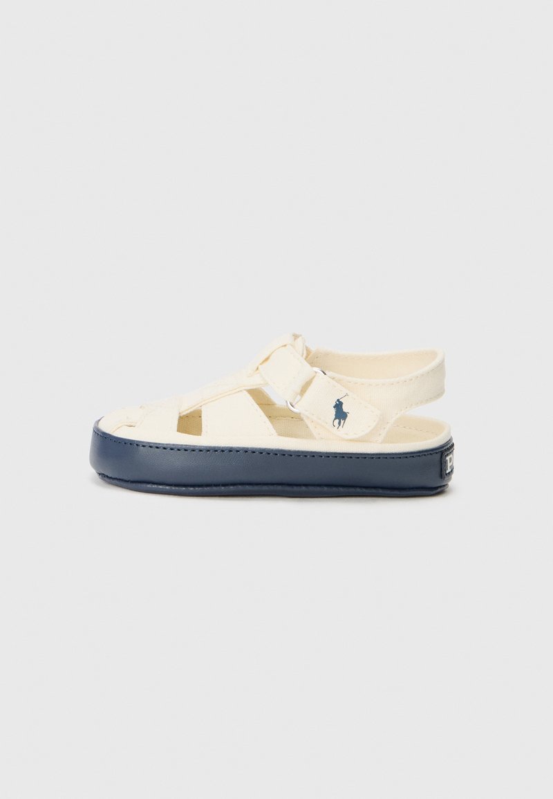 Beige canvas sandaal met een marineblauwe rubberen zool en een verstelbare band. Heeft een uitsnijd ontwerp en een klein logo detail aan de zijkant.