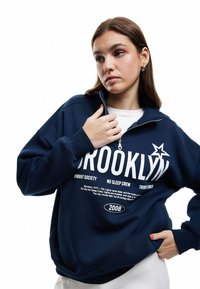 Sudadera con cremallera azul marino con texto gráfico en blanco y negro, que presenta un acento de estrella. Fabricada con un tejido suave y texturizado, con mangas largas.