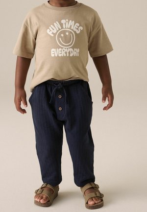 Enfant portant un t-shirt beige avec le texte "Fun Times Everyday" et un visage souriant, un pantalon bleu foncé avec des boutons, et des sandales beiges.