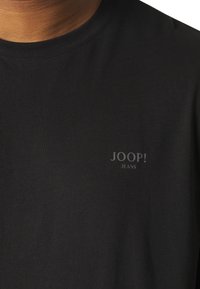 Černé tričko vyrobené z hladkého materiálu, s jemným šedým logem "JOOP! JEANS" vytištěným na levé straně hrudi.