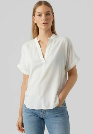 Blouse blanche à manches courtes avec un col en V et des poignets retroussés, fabriquée en tissu léger. L'ourlet est droit et elle a une coupe décontractée.