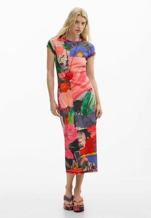 Kvinde står iført en farverig midi-kjole med blomsterprint og korte ærmer, kombineret med platformsandaler og et rødt hjertevedhængs halskæde.