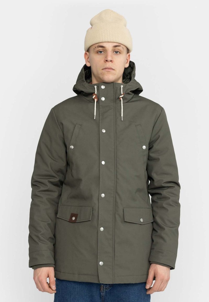 Olivgrüne wasserdichte Jacke mit Kapuze, Druckknöpfen, Vordertaschen und gerippten Bündchen. Trägt eine beigefarbene Strickmütze.