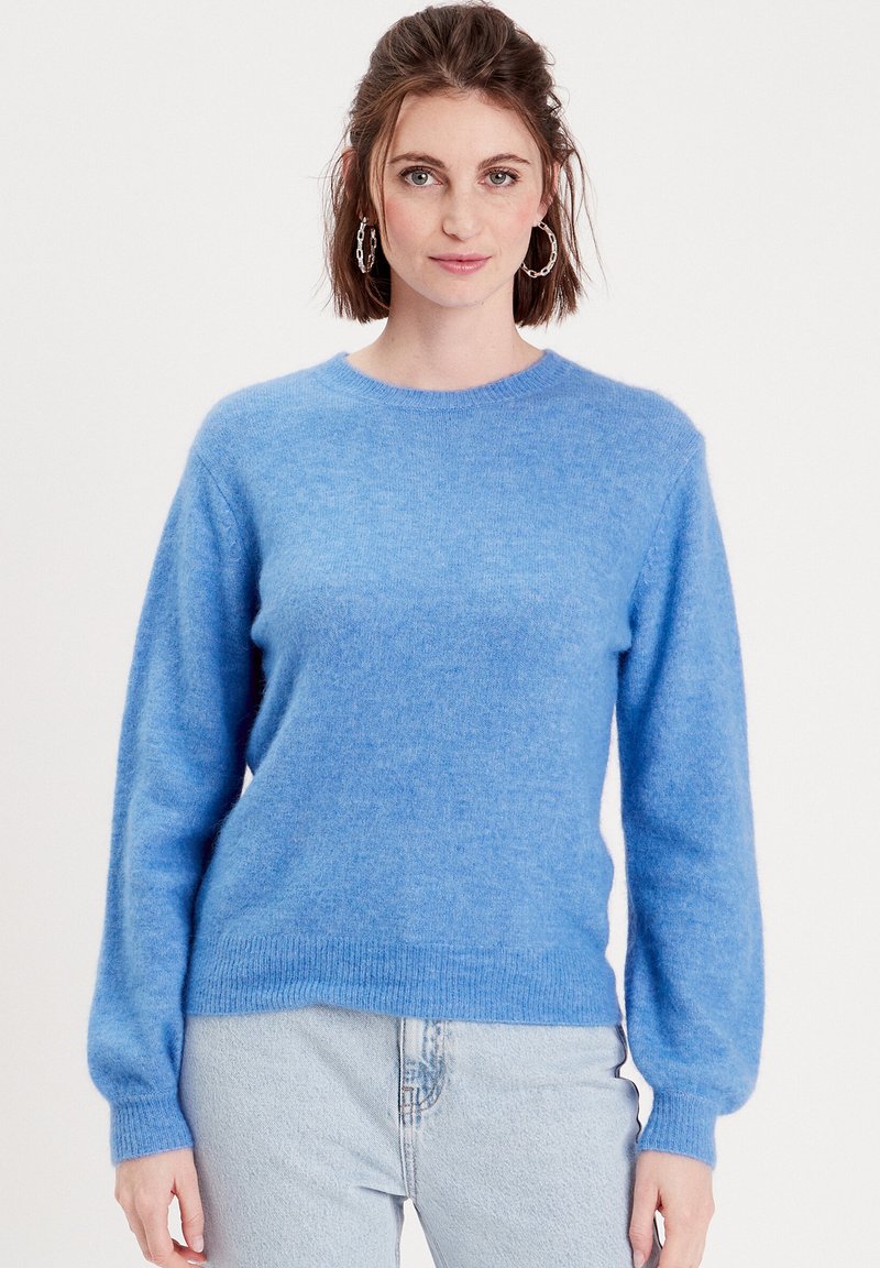Maglione lavorato a maglia di un azzurro chiaro, realizzato in materiale morbido e peloso. Presenta un collo girocollo, maniche lunghe e orlo a costine. Indossato con jeans di jeans chiari.