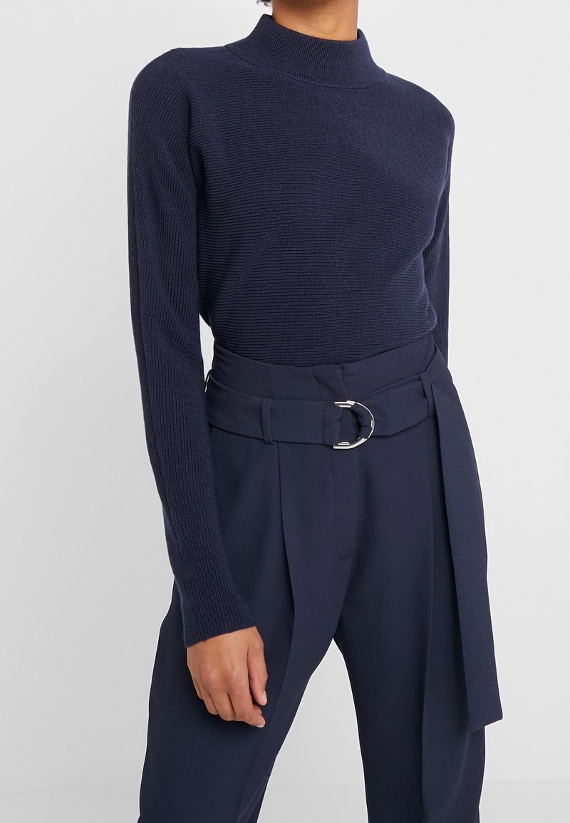Granatowy sweter z ribbowanym splotem i wysokim kołnierzem, w połączeniu z dopasowanymi spodniami o wysokim stanie z paskiem w talii i okrągłą klamrą.