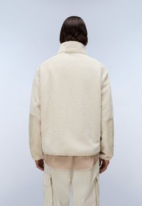 Jachetă din fleece în alb off cu o finisare texturată și un guler înalt, combinată cu un top stratificat bej și pantaloni de culori deschise.