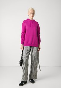 Felpa fucsia con tasca a marsupio, abbinata a pantaloni cargo grigi con tasche e cuciture. Sneakers nere completano l'outfit.