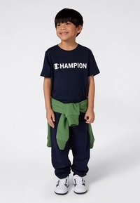 Αγόρι με χαμόγελο, φορώντας ένα ναυτικό μπλουζάκι της Champion, ταιριαστά παντελόνια, άσπρα αθλητικά παπούτσια, και με πράσινη ζακέτα δεμένη στη μέση, που στέκεται σε λευκό φόντο.