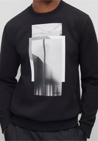 Černá mikina s grafickým designem, který obsahuje vrstvené černobílé fotografie a pruhy. Materiál vypadá měkce a texturovaně.