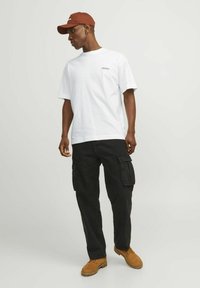 T-shirt bianco in cotone con una piccola grafica, pantaloni cargo neri, cappello marrone e stivali da lavoro beige, che mostrano un outfit casual.