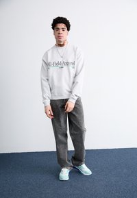Világosszürke pulóver fekete felirattal, laza illeszkedésű szürke farmernadrág, és türkizkék-fehér sneaker cipők kék szőnyegen. Egyszerű nyaklánc kiegészítő.