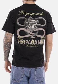 Uomo che indossa una maglietta nera con un serpente attorcigliato intorno a una testa femminile classica e testo "Propaganda" e "C'è solo denaro" sul retro.