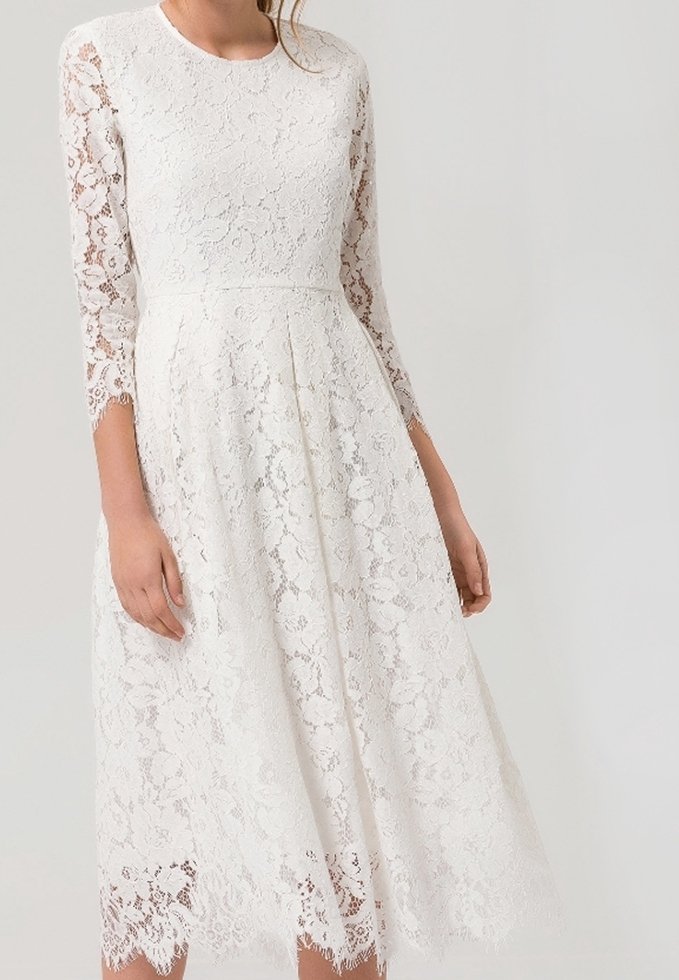 Bílé krajkové šaty s dlouhými rukávy, upnutým živůtkem a rozšířenou sukní. Mají vlnitý lem a květinový krajkový vzor, vyrobené z lehkého materiálu.