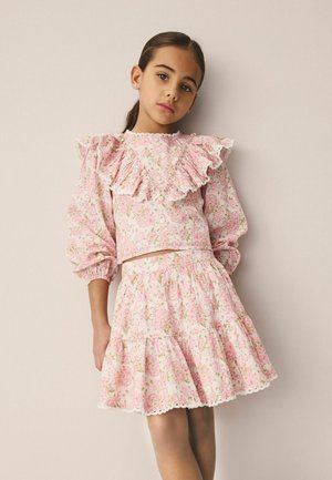 Junges Mädchen mit dunklen Haaren in einem rosa Blumenkleid mit Rüschen an Brust und Rock, stehend vor einem einfarbigen beigen Hintergrund.