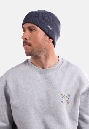 Bonnet en tricot côtelé marine avec une étiquette ; porté avec un sweat-shirt gris clair comportant une broderie florale colorée sur la poitrine.