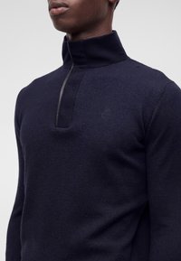 Homme portant un pull zippé bleu marine foncé avec un col montant, montrant la partie supérieure du torse et la partie inférieure de son visage sur un fond uni.
