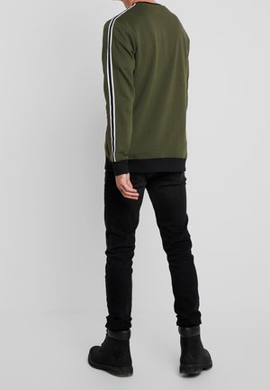 Mann trägt olivgrünes Sweatshirt mit weißen Seitenstreifen, schwarze Jeans und schwarze Stiefel, steht mit dem Rücken zum grauen Hintergrund.