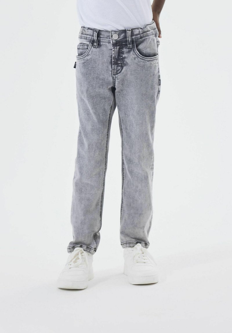 Jeans de mezclilla gris con cinco bolsillos, acabado desgastado, cierre de botón y cremallera, y costuras en contraste. Combinados con zapatillas blancas.