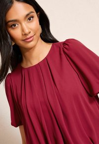 Burgundy-paito lyhythihainen, jossa on pyöreä pääntie ja pystyrypytyksiä, valmistettu sileästä ja kevyestä kankaasta.