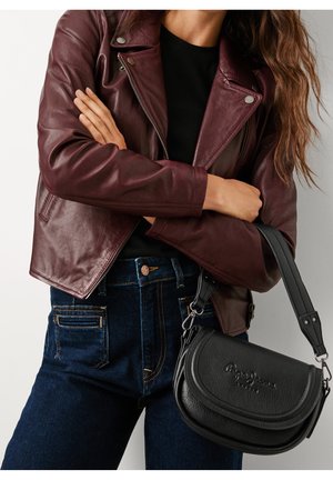 Mujer que lleva una chaqueta de cuero granate y vaqueros azul oscuro, sosteniendo un bolso bandolera negro con el logo de la marca estampado.