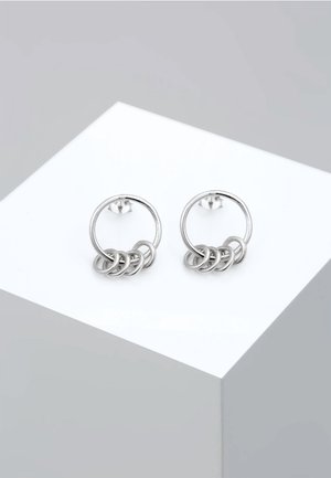 Boucles d'oreilles créoles argentées, chacune composée de quatre petites anneaux entrelacés, présentées sur un socle blanc sur un fond gris clair.
