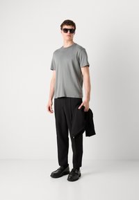 Tricou gri cu mânecă scurtă, pantaloni negri texturați și pantofi negri slip-on. Ținând o jachetă neagră, purtând ochelari de soare închiși la culoare.