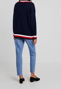 Marineblauer Strickpullover mit rot-weißer und blauer gestreifter Saum- und Kragenpartie, kombiniert mit hellblauen tapered Jeans und schwarzen Slippern.