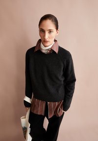 Pull en tricot gris foncé sur une chemise rayée marron et blanche. Pantalon noir et un sac fourre-tout clair, avec un fond à texture subtile.