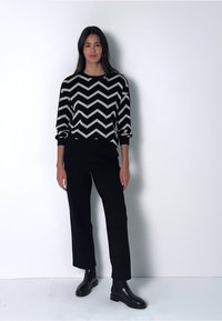 Maglione bianco e nero con motivo a zigzag e maniche lunghe, abbinato a pantaloni a palazzo neri e stivali alla caviglia. Sfondo semplice.