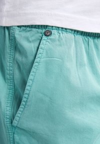 Shorts turquoise clair en tissu doux, avec une taille à cordon de serrage, une poche latérale et un petit détail en métal sur le bouton.
