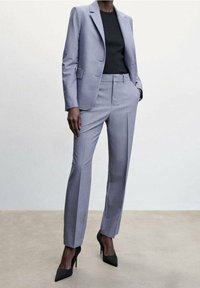 Costume gris clair sur mesure avec une veste structurée à revers crantés et un pantalon slim. Complété par des talons noirs.