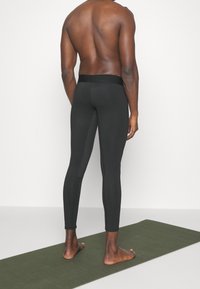 Leggings deportivos negros con un diseño ajustado, textura suave y una cinturilla ancha, exhibidos desde una vista trasera sobre una alfombra verde.