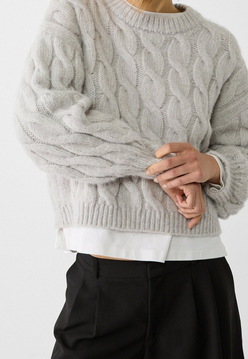 Hellegrauer Zopfmuster-Pullover mit hohem Kragen, strukturiertem Muster und Rippensaum, über einem weißen Hemd getragen und kombiniert mit schwarzen, taillierten Hosen.