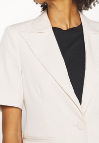 Světle béžový blazer s klopovým límcem, krátkými rukávy, jedním knoflíkem a jemnou texturou, spárovaný s černou košilí.