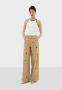 White tank top with green trim, paired with wide-leg tan pants featuring blue flower embroidery and the word "SOL" on the front. Silver shoes.