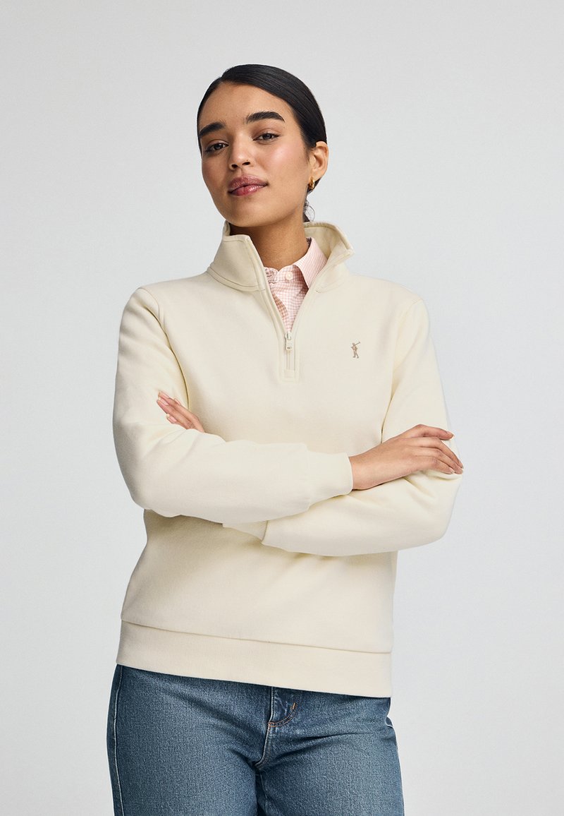 Sudadera de color beige claro con cuello de media cremallera, puños acanalados y un pequeño logo en el pecho, combinada con jeans de mezclilla.
