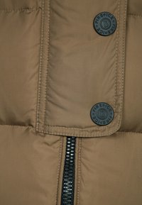 Detailní záběr na hnědou zateplenou bundu s černým zipem a dvěma černými knoflíky s vyraženým logem "FIRE + ICE".