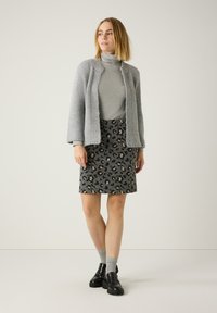 Grau gestrickte Strickjacke über einem grauen Rollkragenpullover, kombiniert mit einem grauen Rock im Leopardenmuster. Das Modell trägt schwarze Slipper und hellgraue Socken.