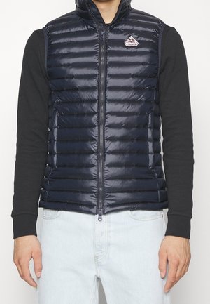 Mann trägt eine marineblaue gesteppte ärmellose Weste über einem schwarzen Langarmhemd und hellblauen Jeans, steht vor einem schlichten Hintergrund.