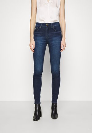 Femme portant un jean skinny bleu foncé, un top blanc sans manches et des bottines noires, debout devant un fond blanc uni.