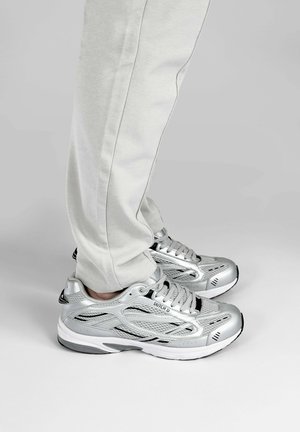 Benedenlijf met lichtgrijze broek en zilveren veterschoenen staand op een grijze vloer met een effen achtergrond.