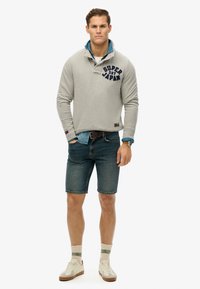 Szary pulower z logo "SUPER DRY JAPAN" nałożony na niebieską koszulę dżinsową; zestawiony z ciemnoniebieskimi szortami dżinsowymi i białymi trampkami.