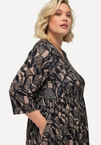 Vrouw met blond haar die een zwart-beige gestreepte jurk met driekwart mouwen en een gouden ketting draagt, met haar hand in haar zak.