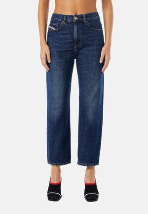 Kobieta w niebieskich, wysokim stanem, prostych dżinsach i czarnych sandałkach na obcasie z czerwonym obszyciem, stojąca na białym tle.