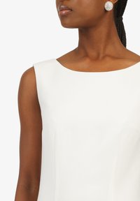 Robe blanche sans manches avec un large décolleté, en tissu texturé et à la coupe structurée. Accompagnée d'une grande boucle d'oreille clou scintillante.