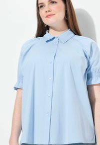 Camicia azzurra chiaro con colletto, maniche a palloncino, chiusura a bottoni e polsini elastici. Tessuto morbido con una vestibilità comoda e senza motivi visibili.