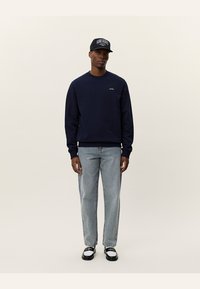 Marineblå genser med rund hals, kombinert med lyseblå denimjeans. Svart og hvite sko, og en mørk caps med tekstlogo.